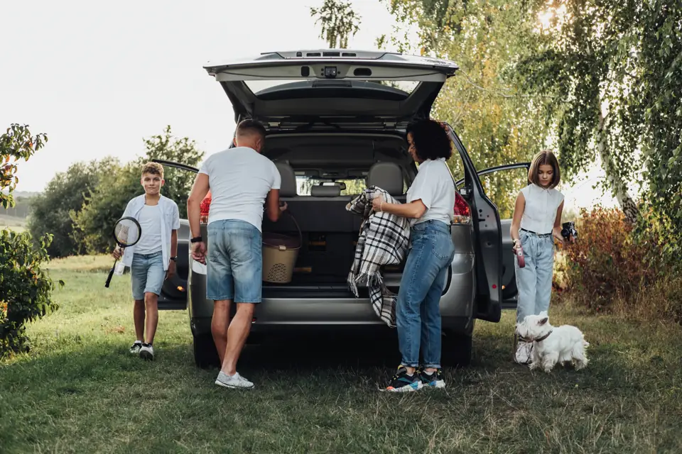 A family loading the back of a car for a trip, their little white dog beside them